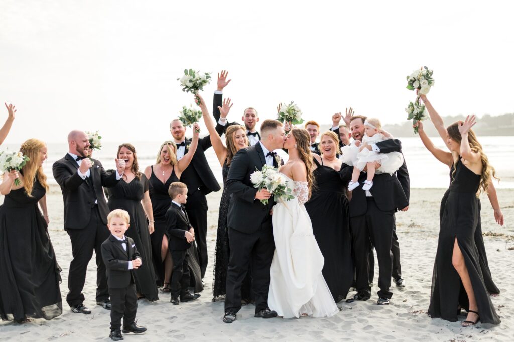 Candid wedding party photo celebrating bride and groom kissing on the beach 
