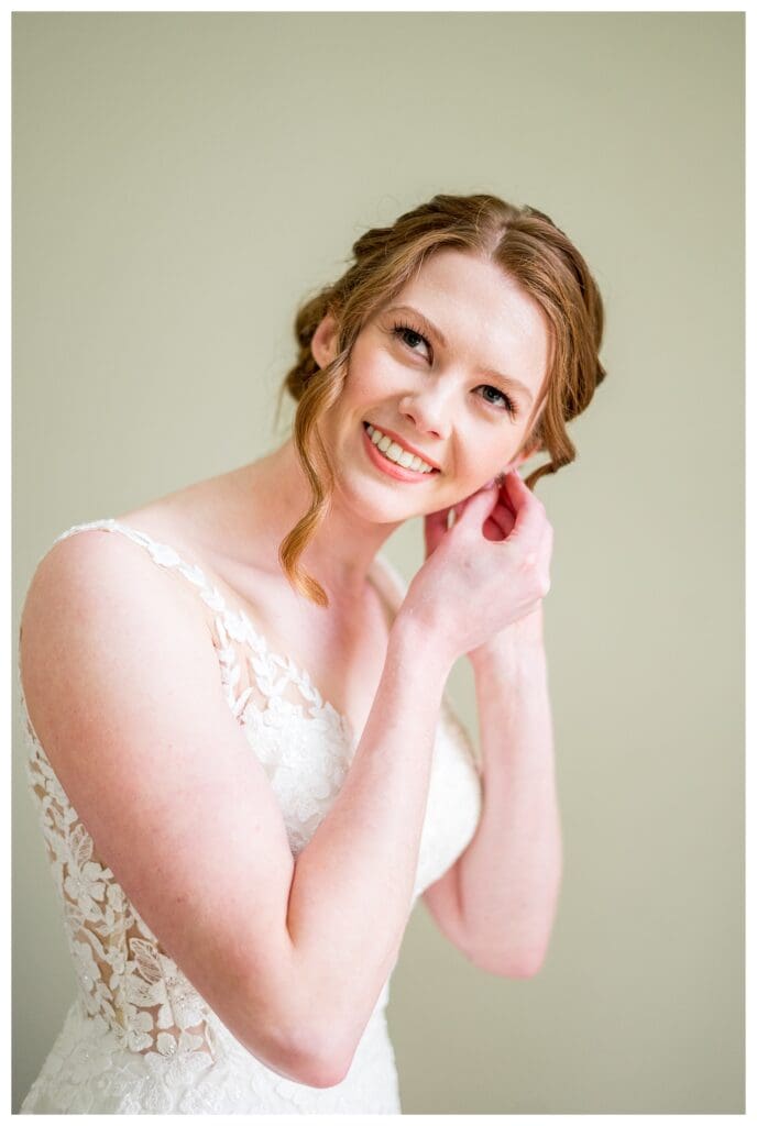 Bride adjusting her earring while getting ready, wearing a lace gown.