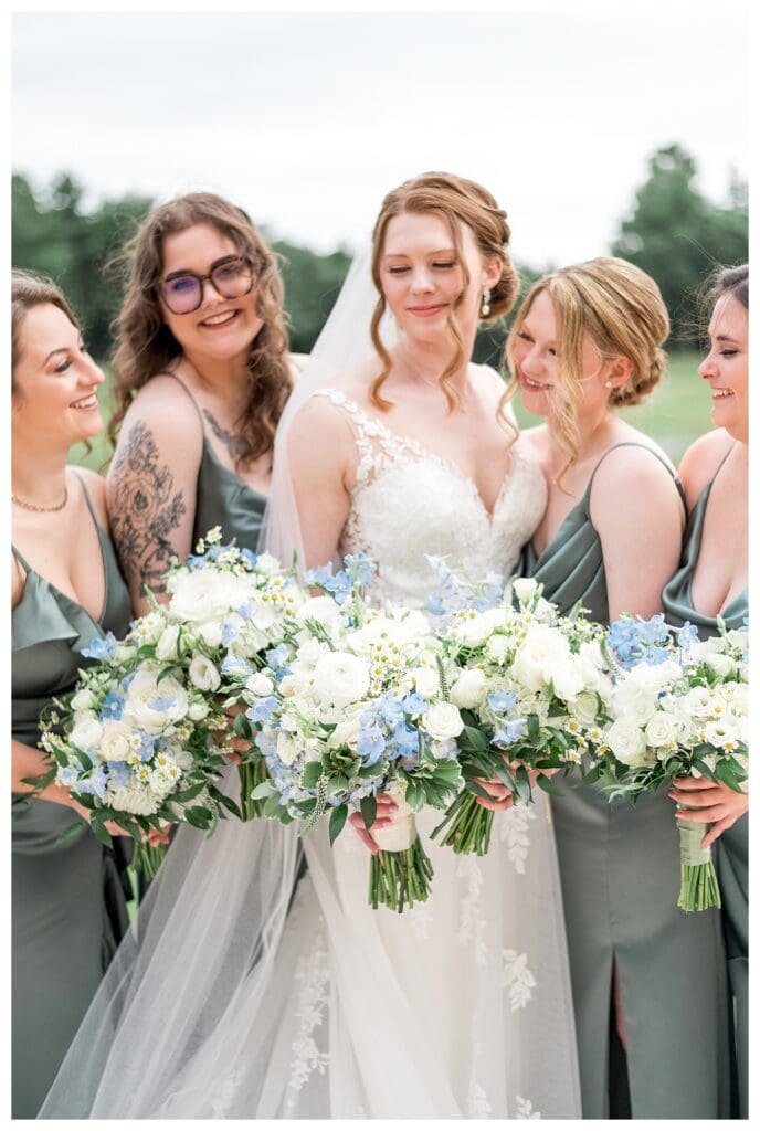 Bridesmaids in sage green dresses hold white and blue floral bouquets.