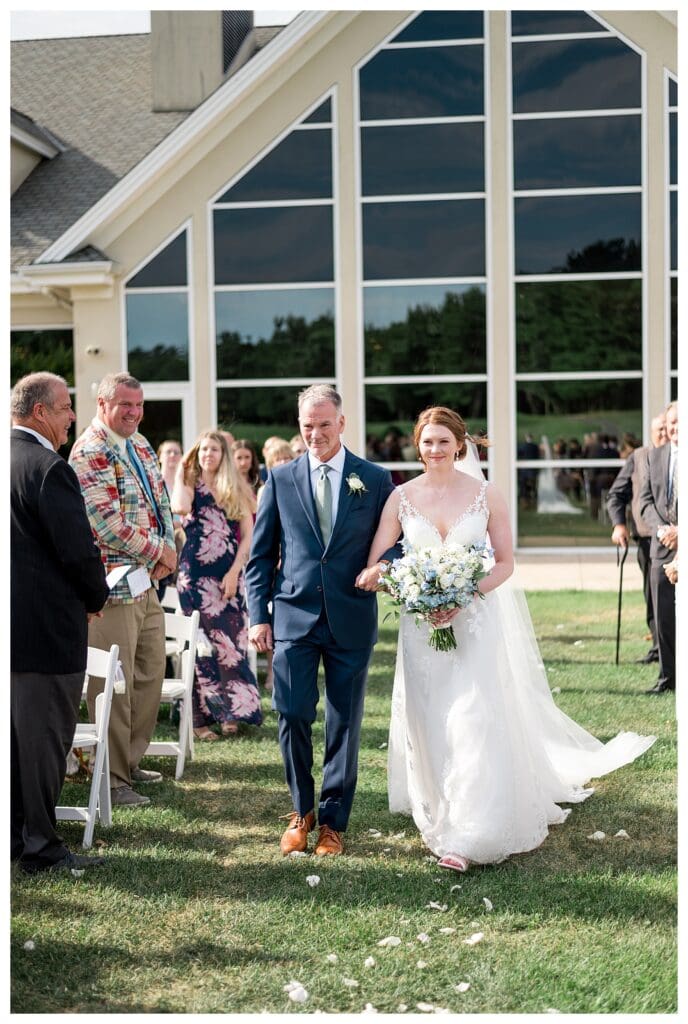 Bride walking down the aisle with her father, smiling at her groom ahead.
