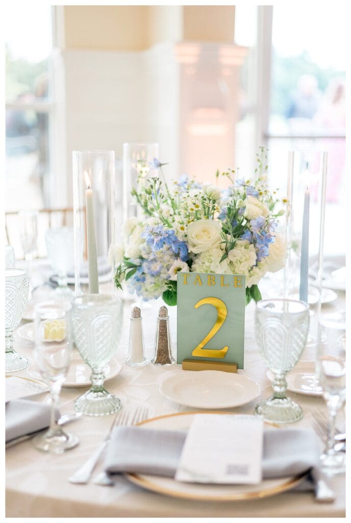 Table setting with gold-accented charger plates, blue linen napkins, and numbered table card.
