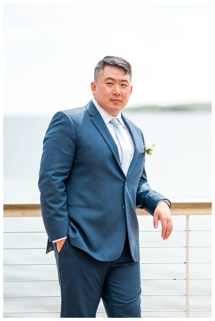 Groom standing confidently in navy suit, boutonniere pinned, with ocean backdrop.