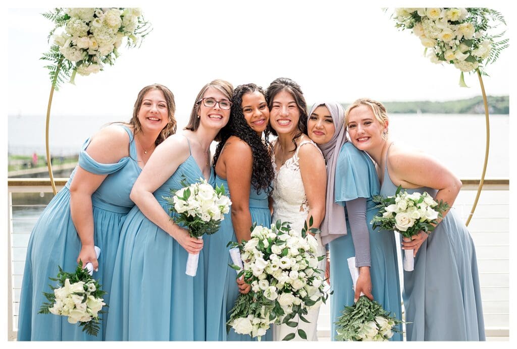 Bridesmaids in coastal blue gowns gathered together, holding white flower bouquets.