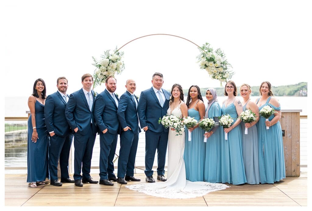 Full wedding party lined up on the deck, dressed in coastal blue attire with white florals.