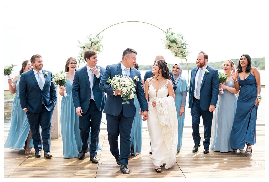 Full wedding party smiling under a circular floral arch at a New England seaside venue.
