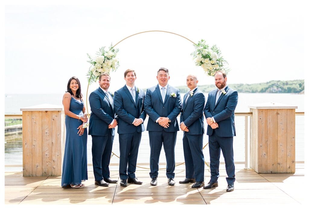 Groomsmen in navy suits smiling together on the seaside deck at a Massachusetts wedding.