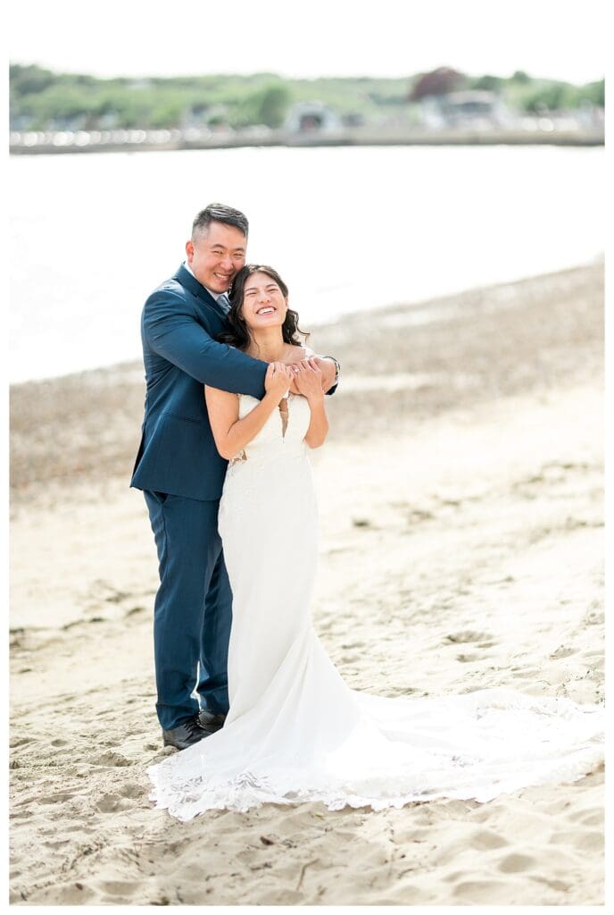 Groom holding bride close with waves in the distance at a Massachusetts coastal wedding.