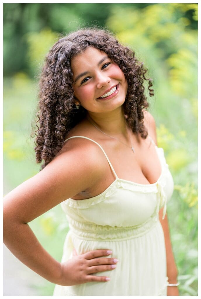 Senior girl in a pale yellow dress posing with one hand on her hip, goldenrod flowers in the background.
