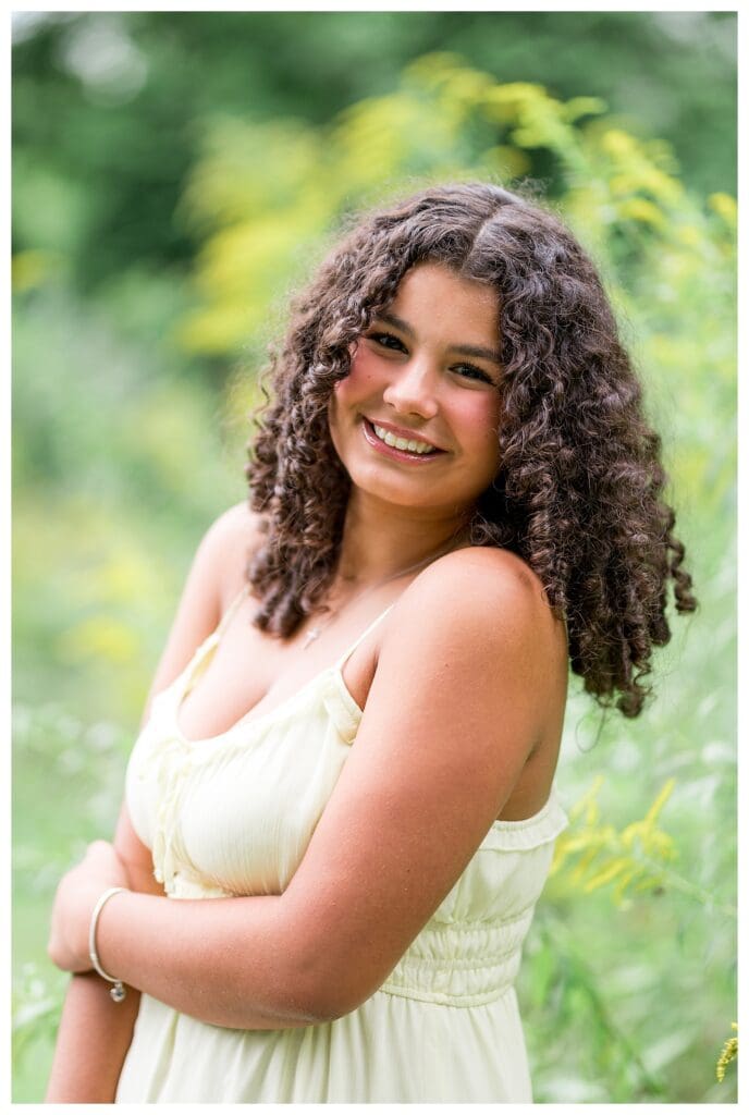 Aubrie smiling brightly in a cream-colored dress surrounded by greenery.