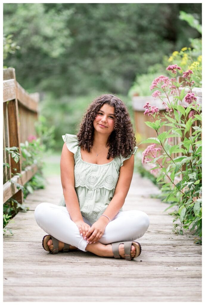 Senior girl in a pale green top, standing on a wooden bridge in a lush outdoor setting.