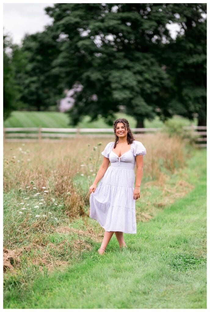 Curly-haired girl in a lavender dress walking through tall grass along a farm fence.