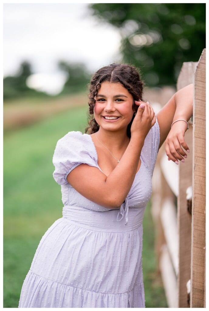 Aubrie resting her elbow on a wooden fence post, smiling brightly during her senior session.
