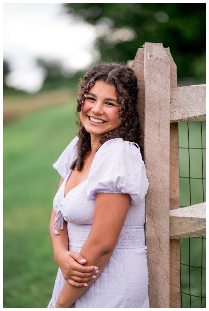 Senior portrait of Aubrie leaning against a rustic fence with her arms crossed, smiling at the camera.