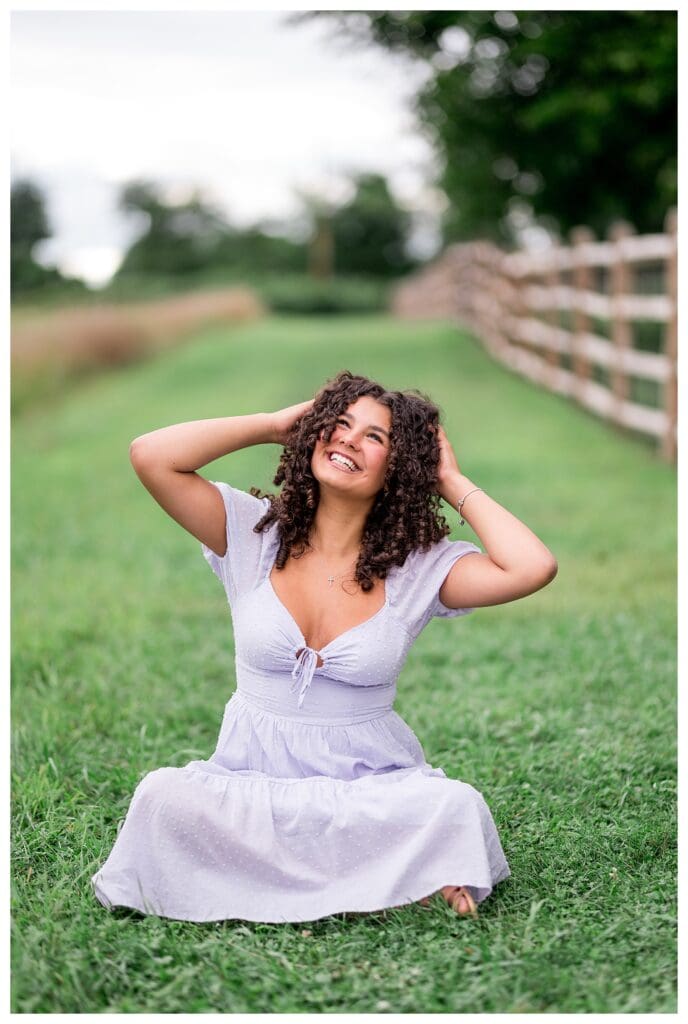 Senior girl in a lavender dress sitting in the grass, looking up with her hands in her curly hair.