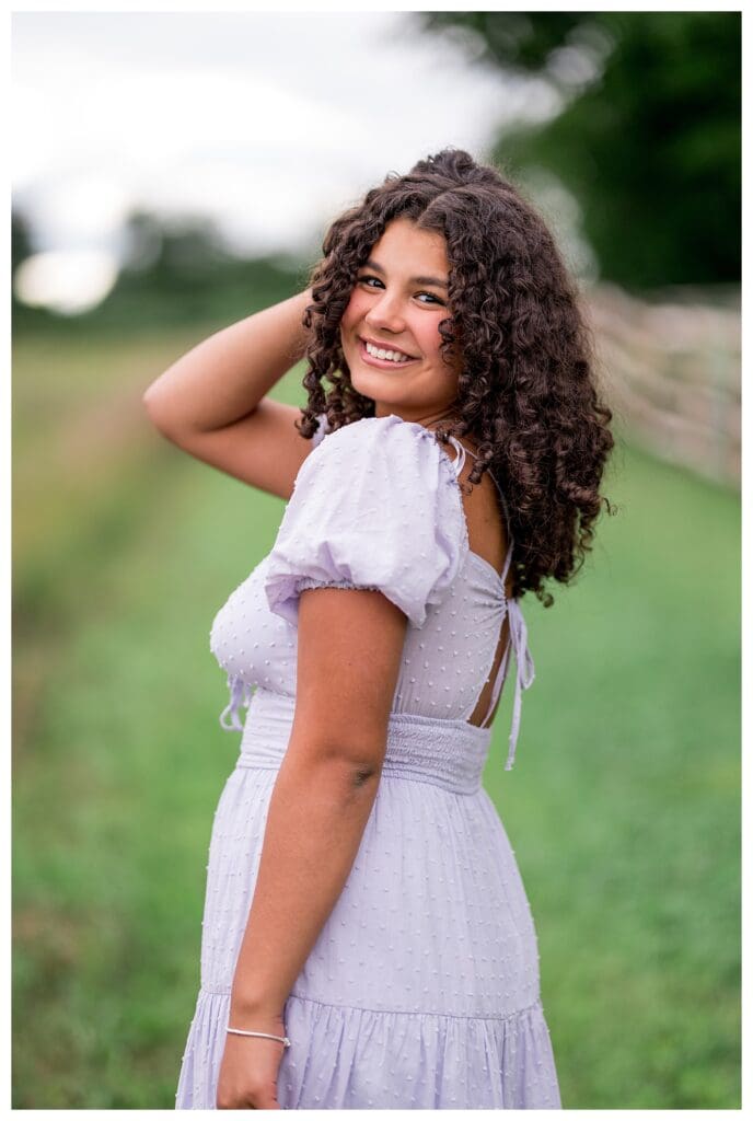 Close-up of a senior girl with curly hair smiling over her shoulder in a lavender dress.