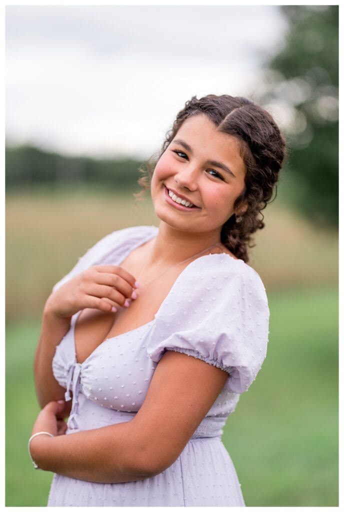 Westford Academy Senior standing in a grassy field wearing a light purple dress, smiling softly at the camera.
