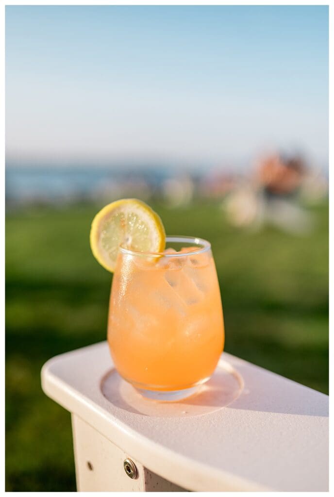 A close-up of the Rose Watermelon Regatta cocktail with a lemon garnish sitting on a white Adirondack chair on the lawn at Castle Hill Inn