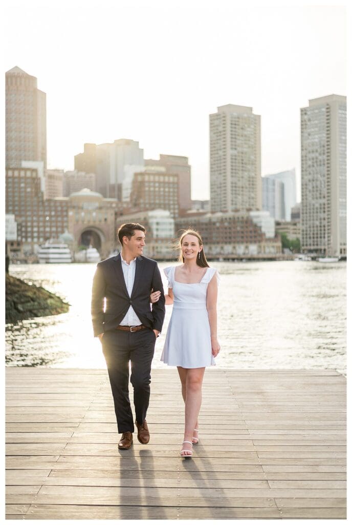 Boston couple walking toward the camera with city reflections on the water.