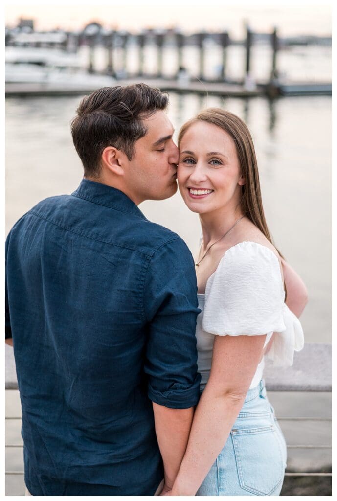 Fiancé gently kissing his partner’s cheek by the pier.