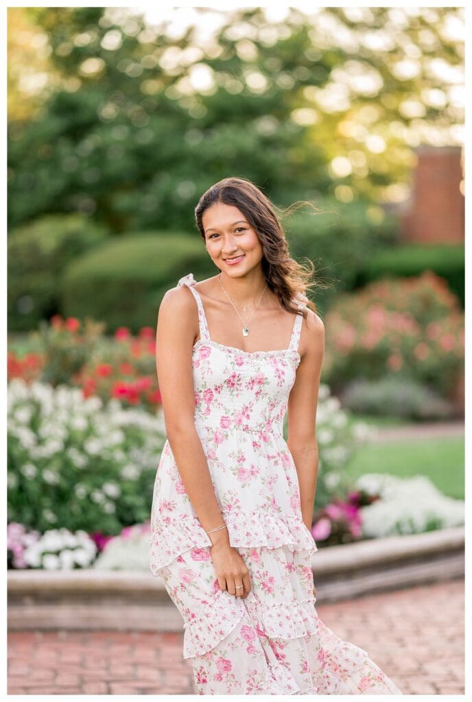 Senior girl smiling in a floral dress surrounded by colorful garden blooms.