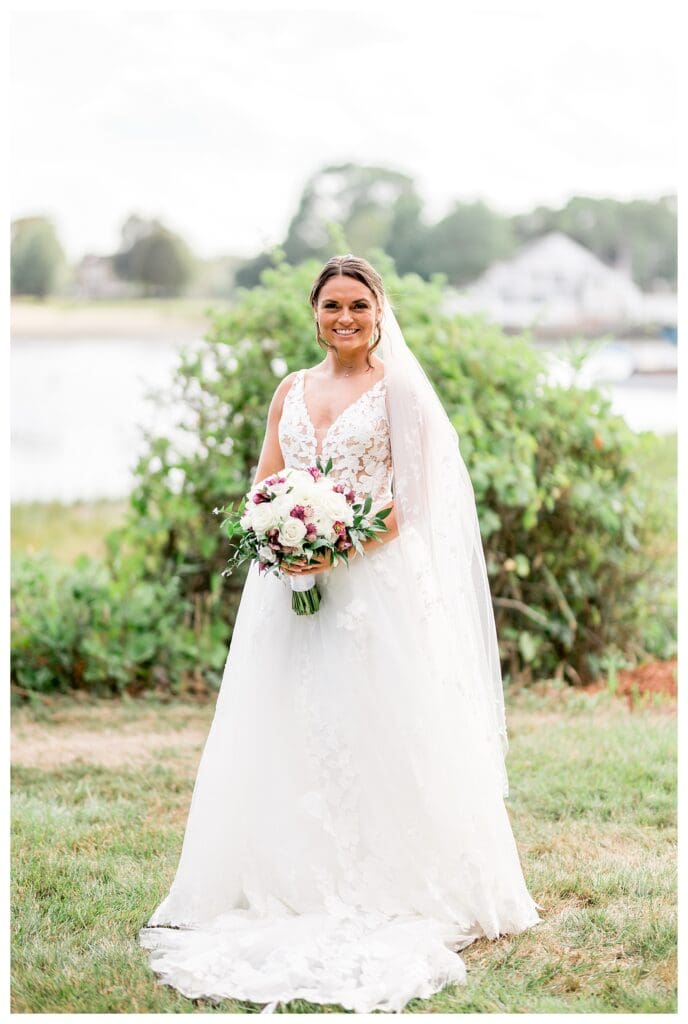 Bride standing by the water holding a white and purple bouquet.