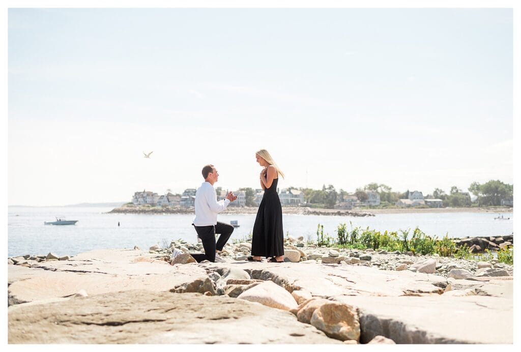 Man down on one knee proposing on coastal jetty with ocean behind them.