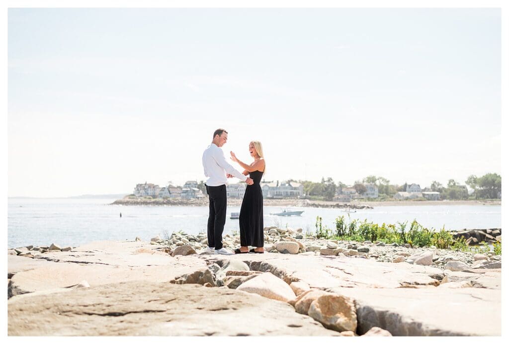 Wide view of proposal on seaside rocks with lighthouse and harbor in distance.