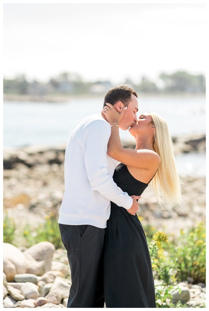 Vertical portrait of engaged couple kissing on rocks along the coast.
