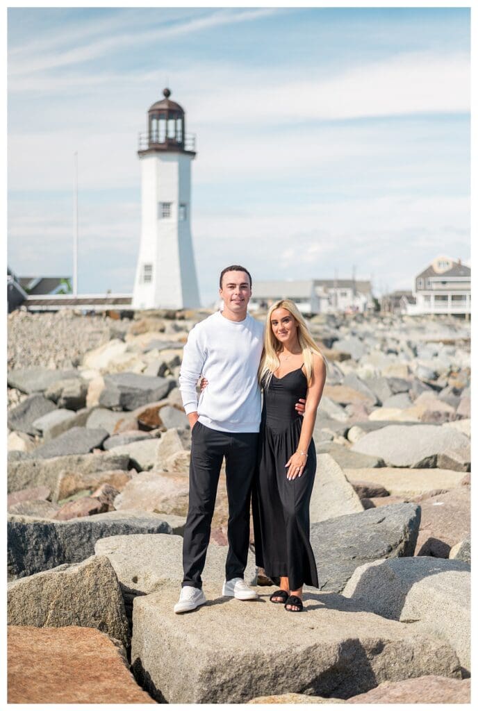 Couple standing on rocky shoreline with Scituate Lighthouse in the background after their proposal.