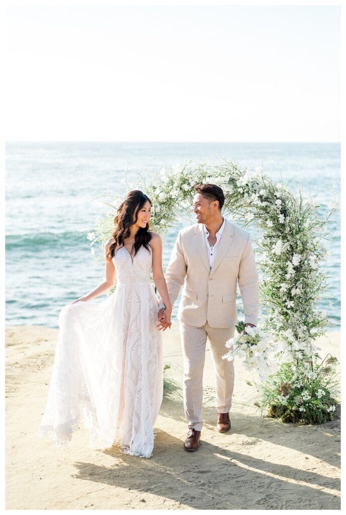 Bride and groom standing together at white floral arch on San Diego beach.