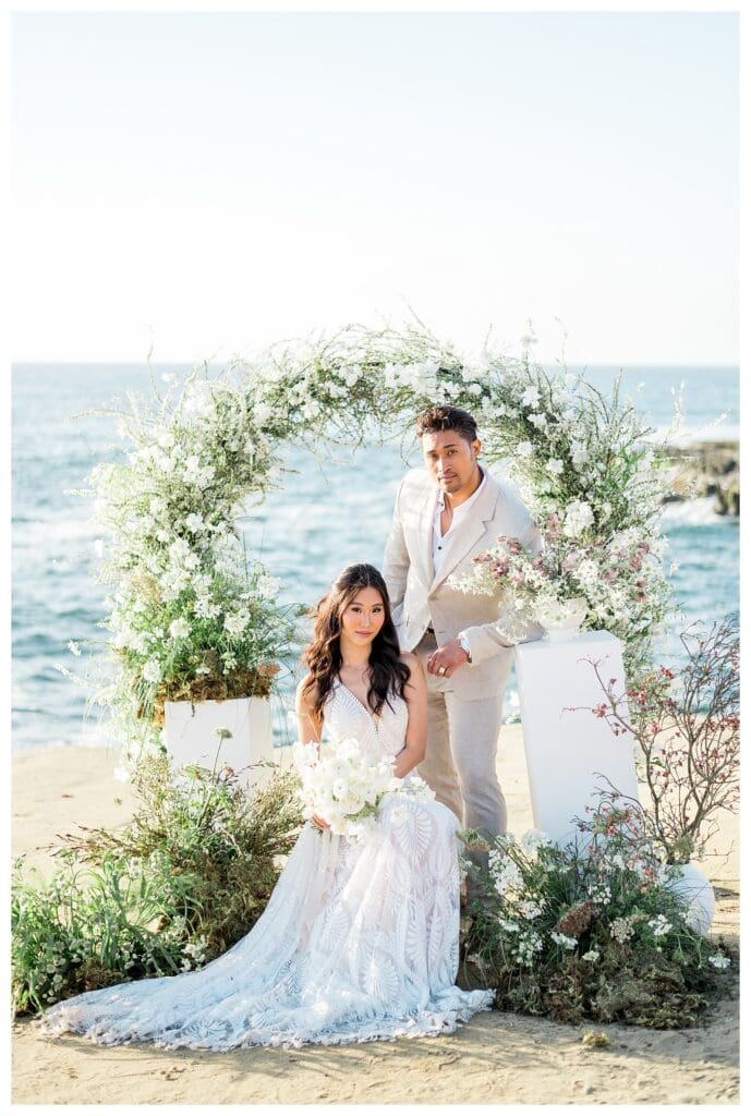 Bride and groom ceremony portrait at white floral arch on San Diego beach.