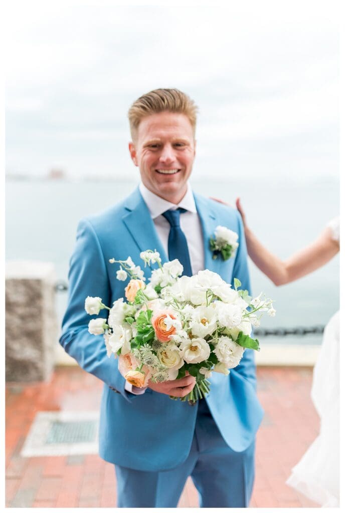 Groom holding bridal bouquet of white and green florals outside Boston Harbor Hotel