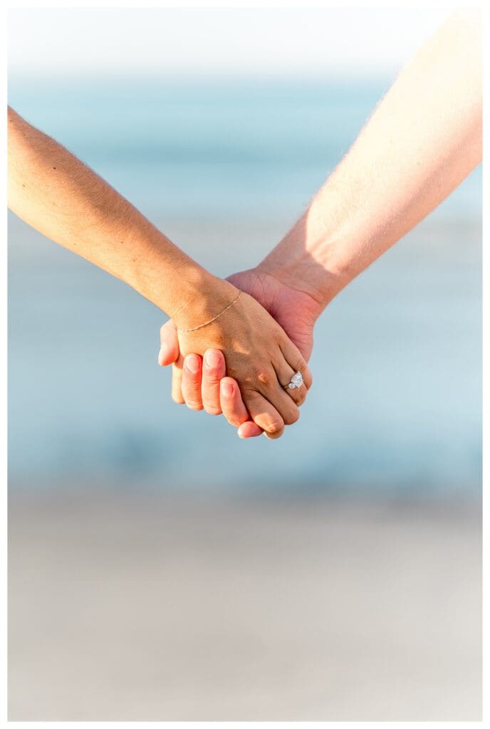 Close-up of hands intertwined on the beach with an engagement ring visible