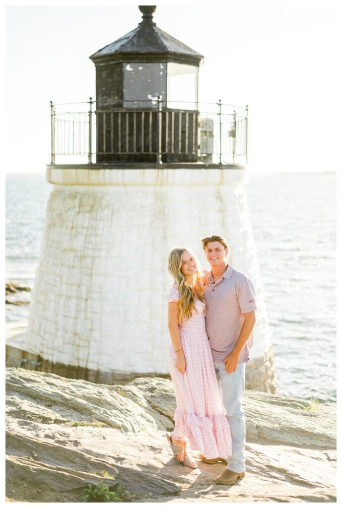 Newly engaged couple stands close together in front of the lighthouse on a bright Newport day.