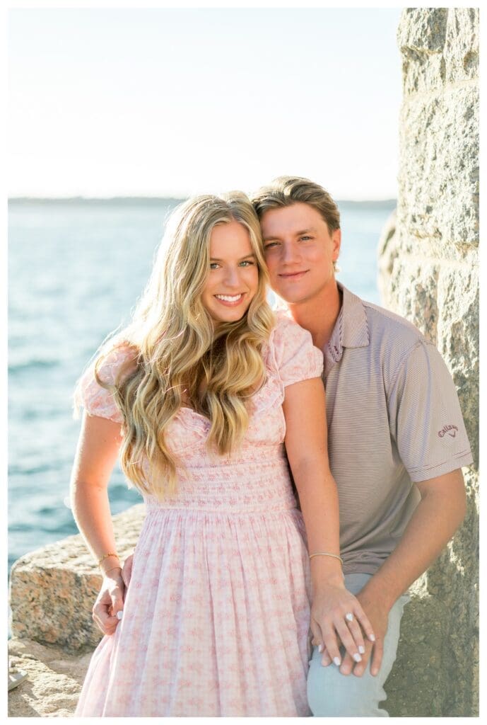 Close portrait of an engaged couple cheek to cheek, smiling, with the ocean and rocks behind them.