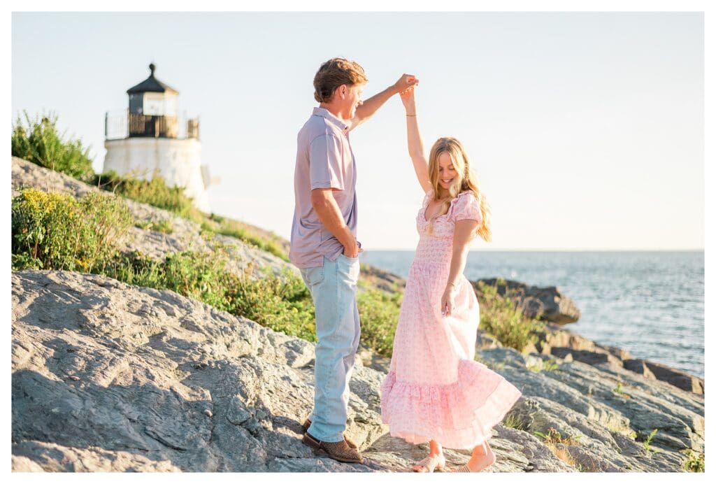 Man twirls his fiancée on the rocks in front of the lighthouse during a Newport engagement session.